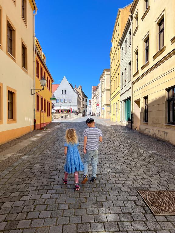 Zwei Kinder, ein Mädchen in blauem Kleid und ein Junge in grauem T-Shirt, gehen auf einer gepflasterten Straße in Torgau mit bunten Häusern und klarem blauem Himmel